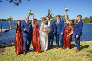 Wedding party celebrating outdoors by lake with bouquets raised in air.