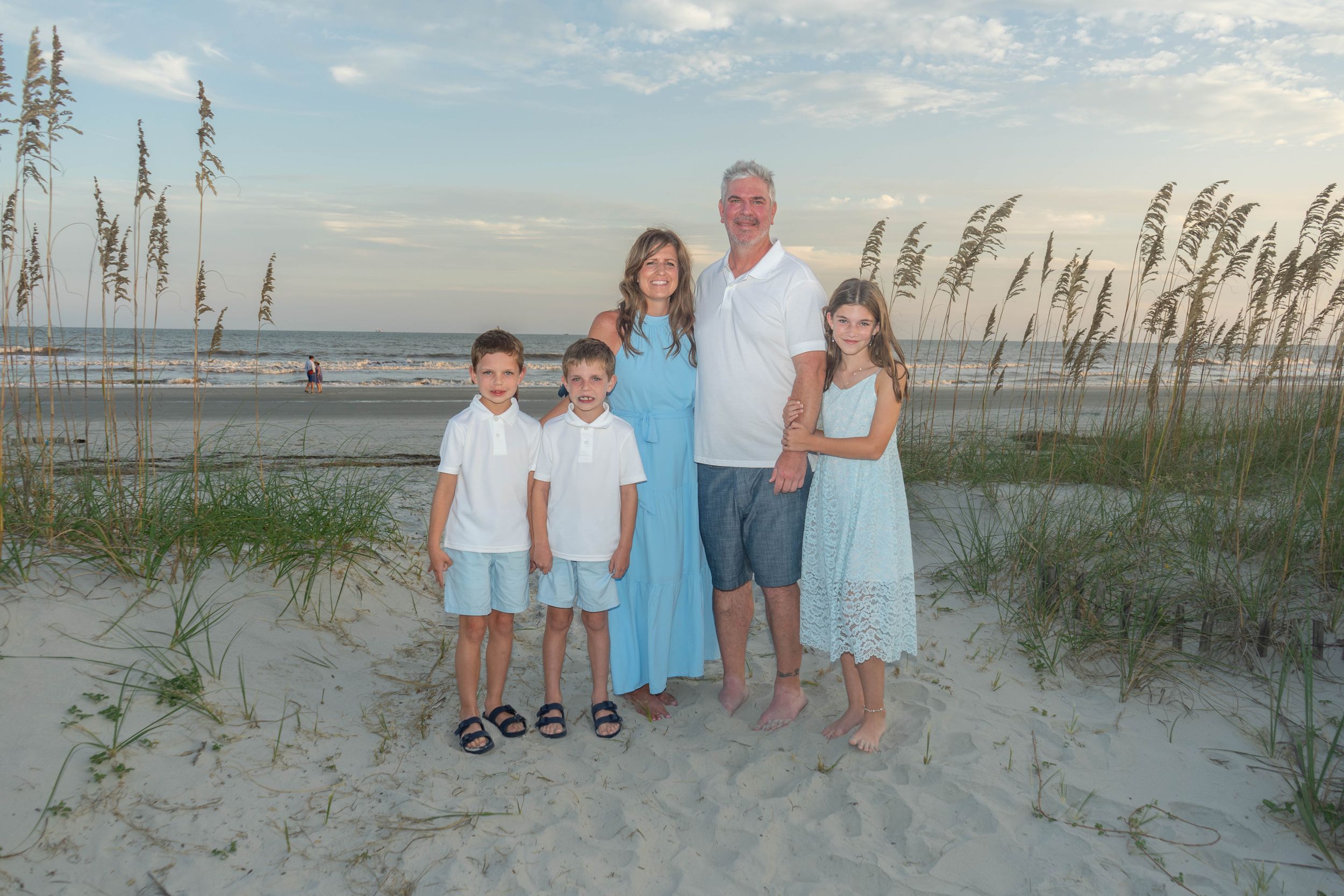 Family of five smiles together on a sandy beach with sea oats and cloudy sky in matching blue attire.