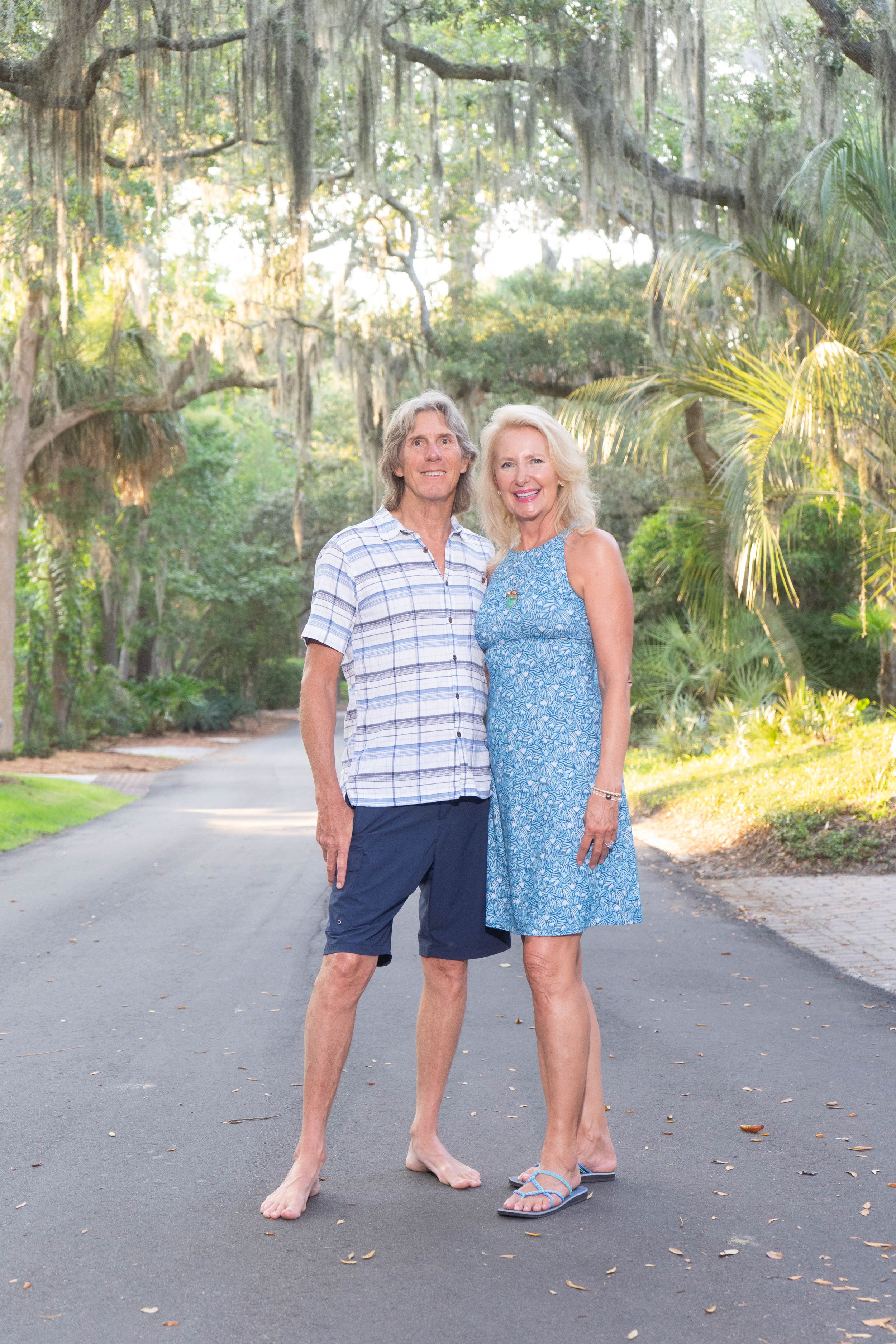 Smiling older couple stands barefoot on a shaded road surrounded by moss-draped trees in warm sunlight.