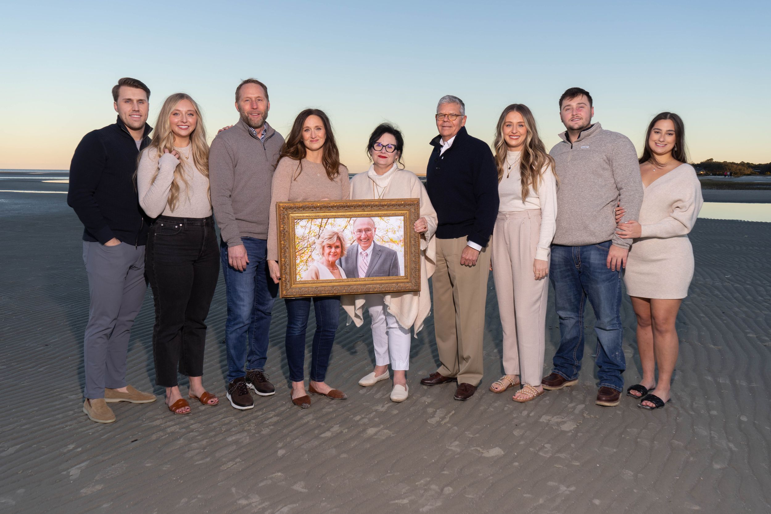Large family group poses on a beach at sunset holding a framed portrait photo of an older couple.