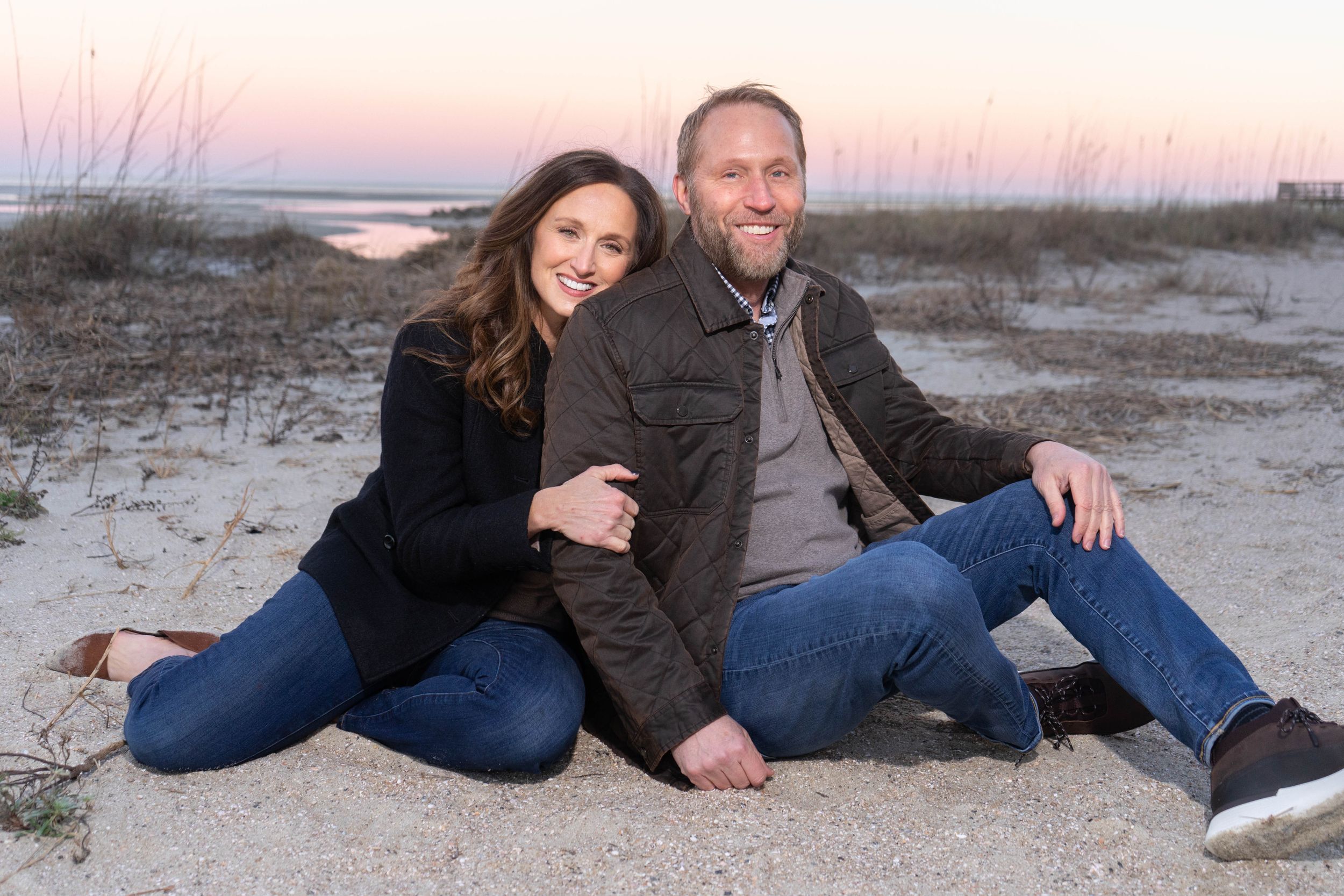 Smiling couple sits together on a sandy beach at dusk, dressed casually in jeans and jackets.