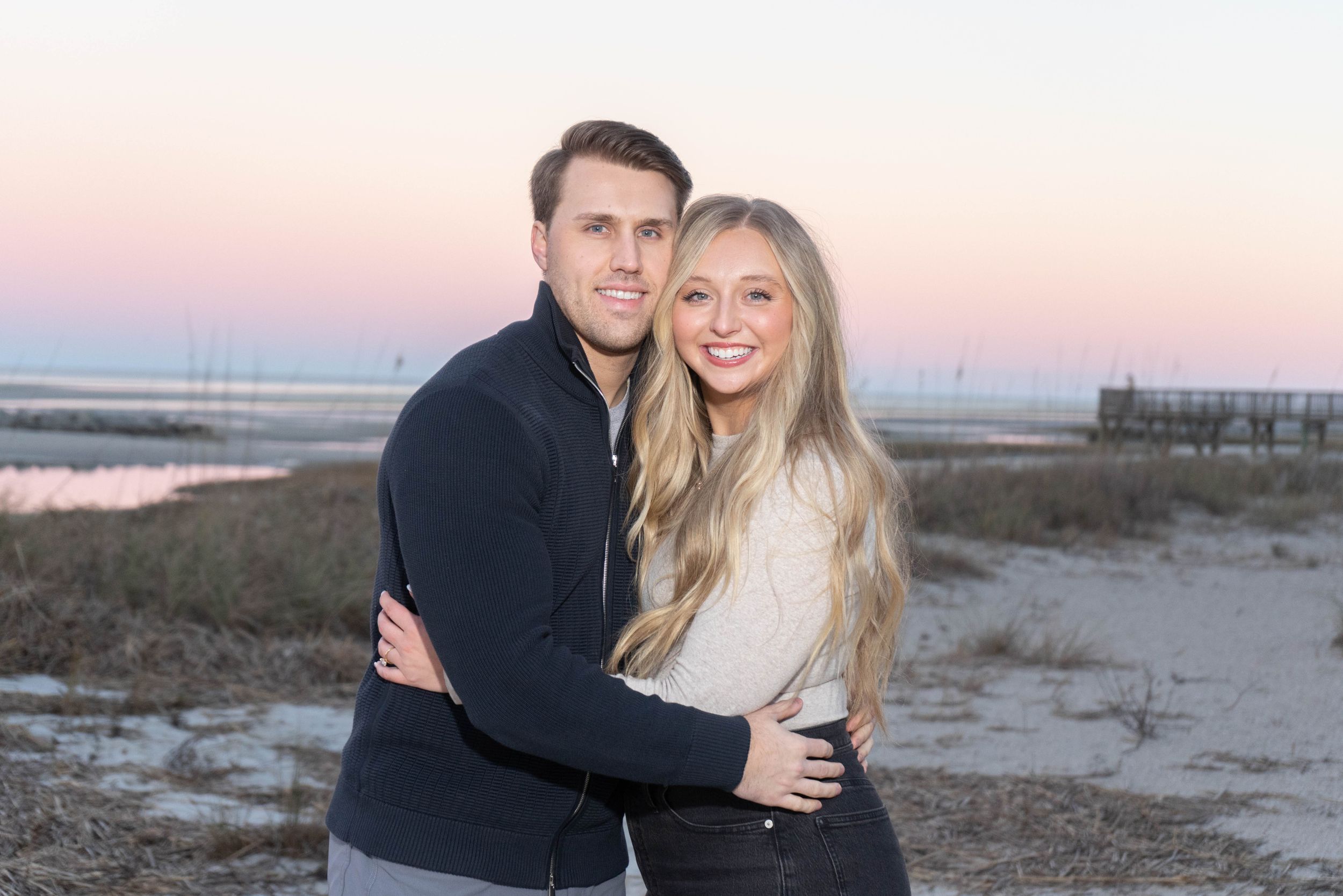 Young couple embraces on a sandy beach at sunset with a colorful pink and blue sky in the background.