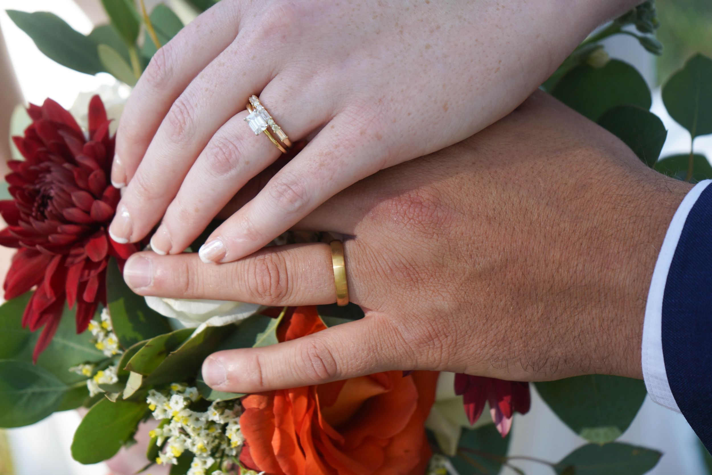 Close-up of newlyweds hands showing wedding rings resting on a bridal bouquet with red and orange flowers.