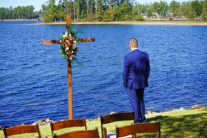 Groom in blue suit standing before floral cross by blue lake.