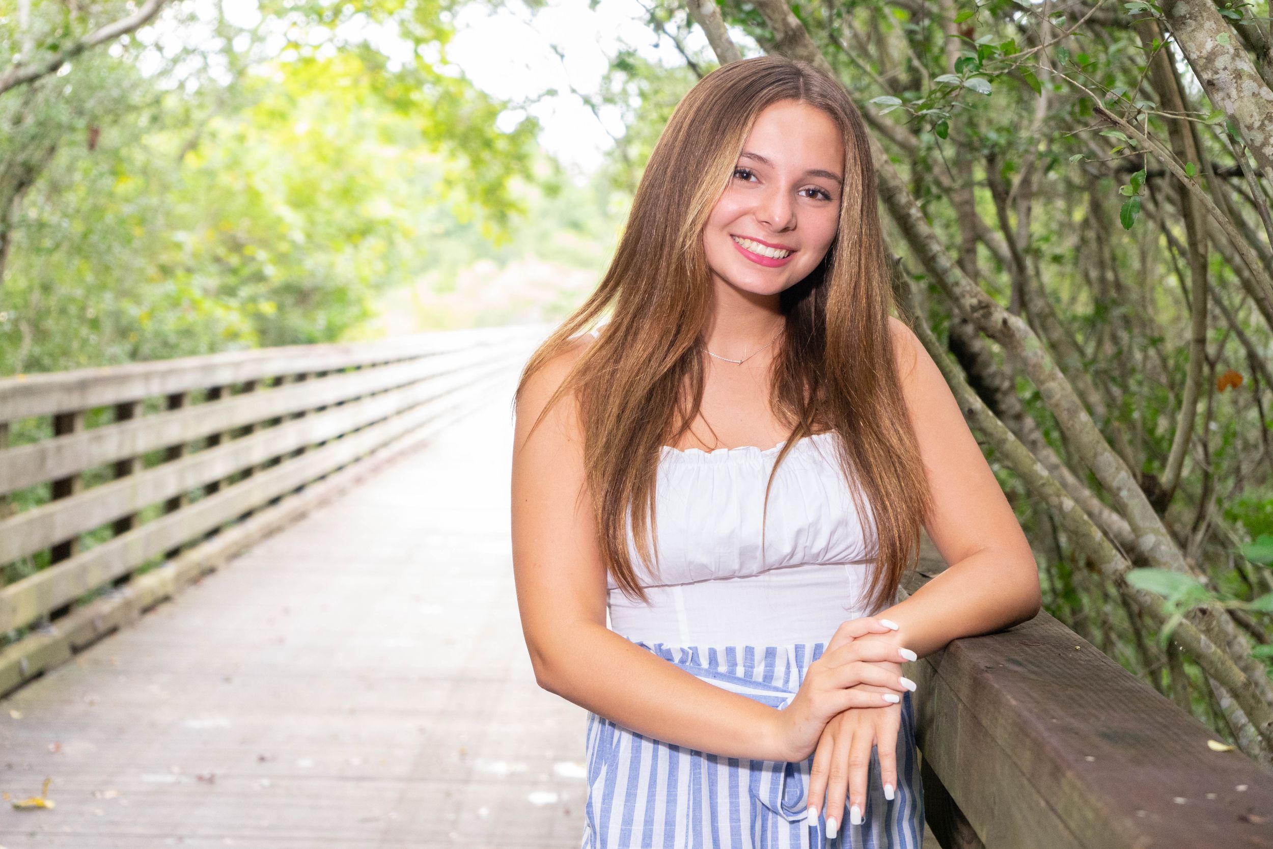 Smiling teenage girl with long brown hair poses on a wooden boardwalk surrounded by lush greenery.