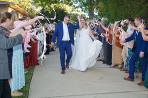 Bride and groom walk through ribbon wand send-off tunnel with cheering wedding guests lining the path.