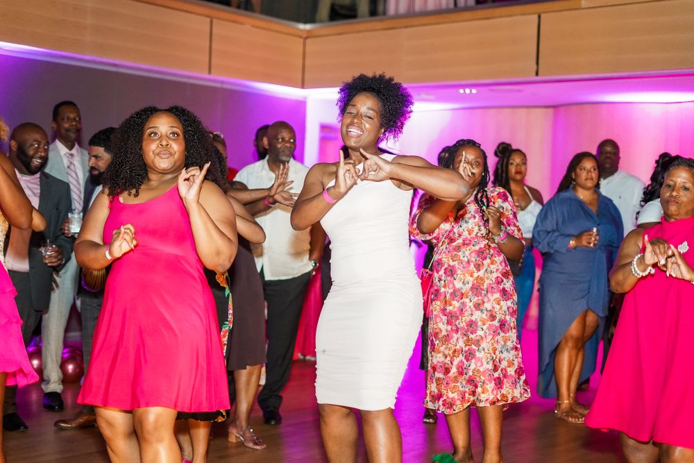 Large group dancing and celebrating in a venue decorated with pink lighting and floral accents.