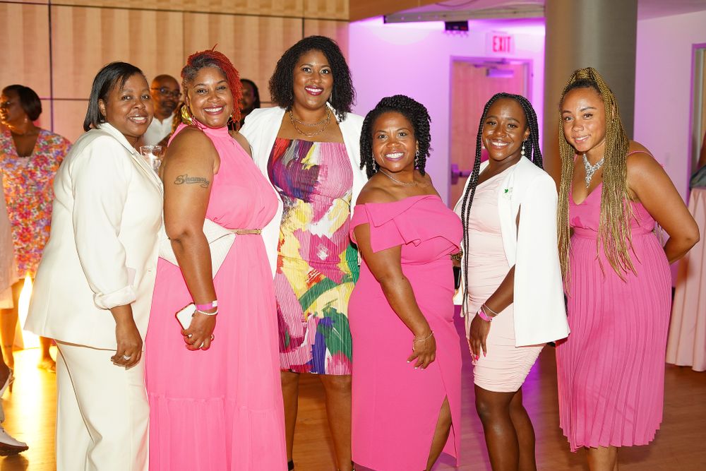 Event attendees in pink and white outfits posing together in a banquet hall with pink ambient lighting.