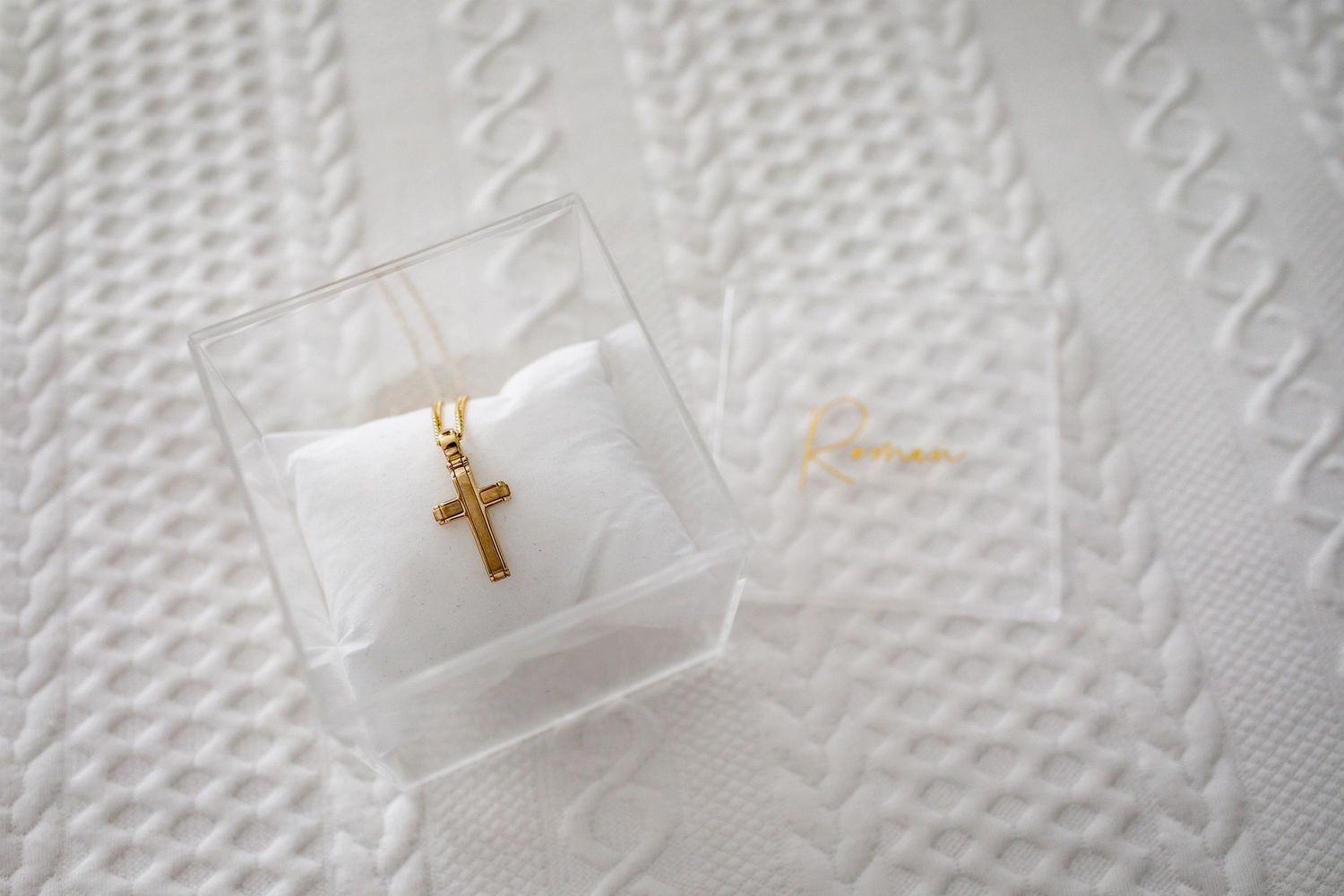 Small gold cross necklace displayed in clear box on white textured background.