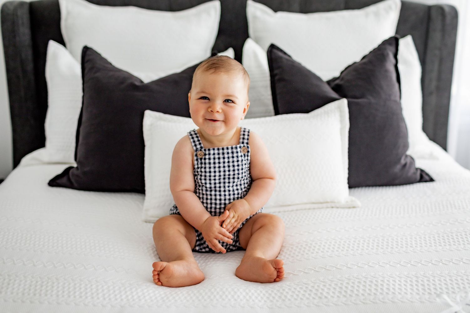 A baby in gingham outfit sitting on a bed with black and white pillows.