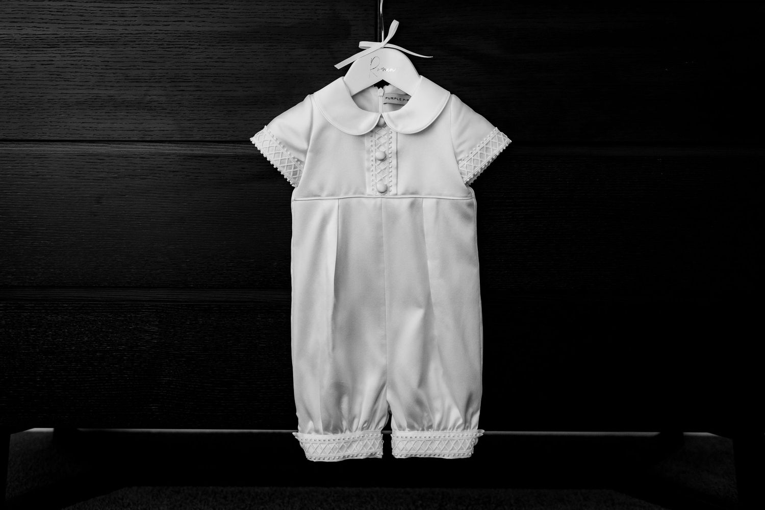 A white christening romper hanging against a dark background.