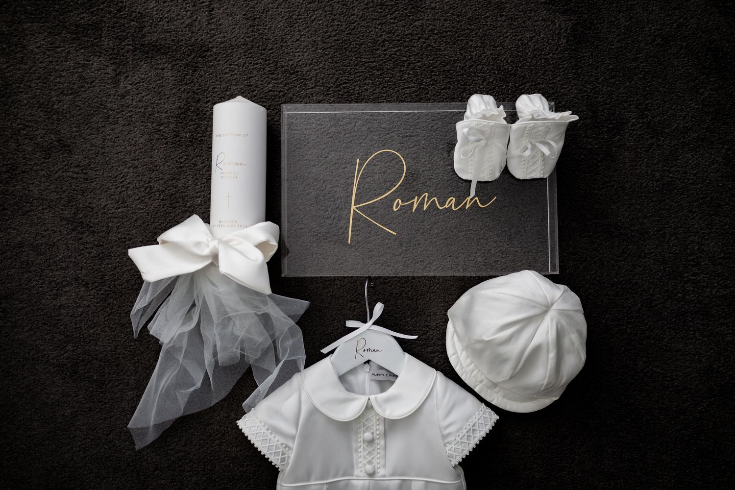 White baptismal outfit with candle, cross necklace, and small shoes arranged on dark background.