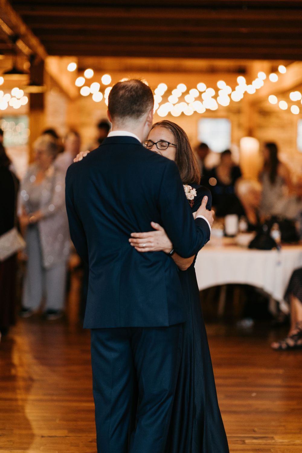 Mother and son share emotional dance at wedding reception with warm string lights overhead.