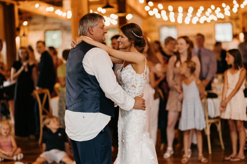Bride in lace gown shares a father-daughter dance at a rustic barn wedding reception with guests watching.