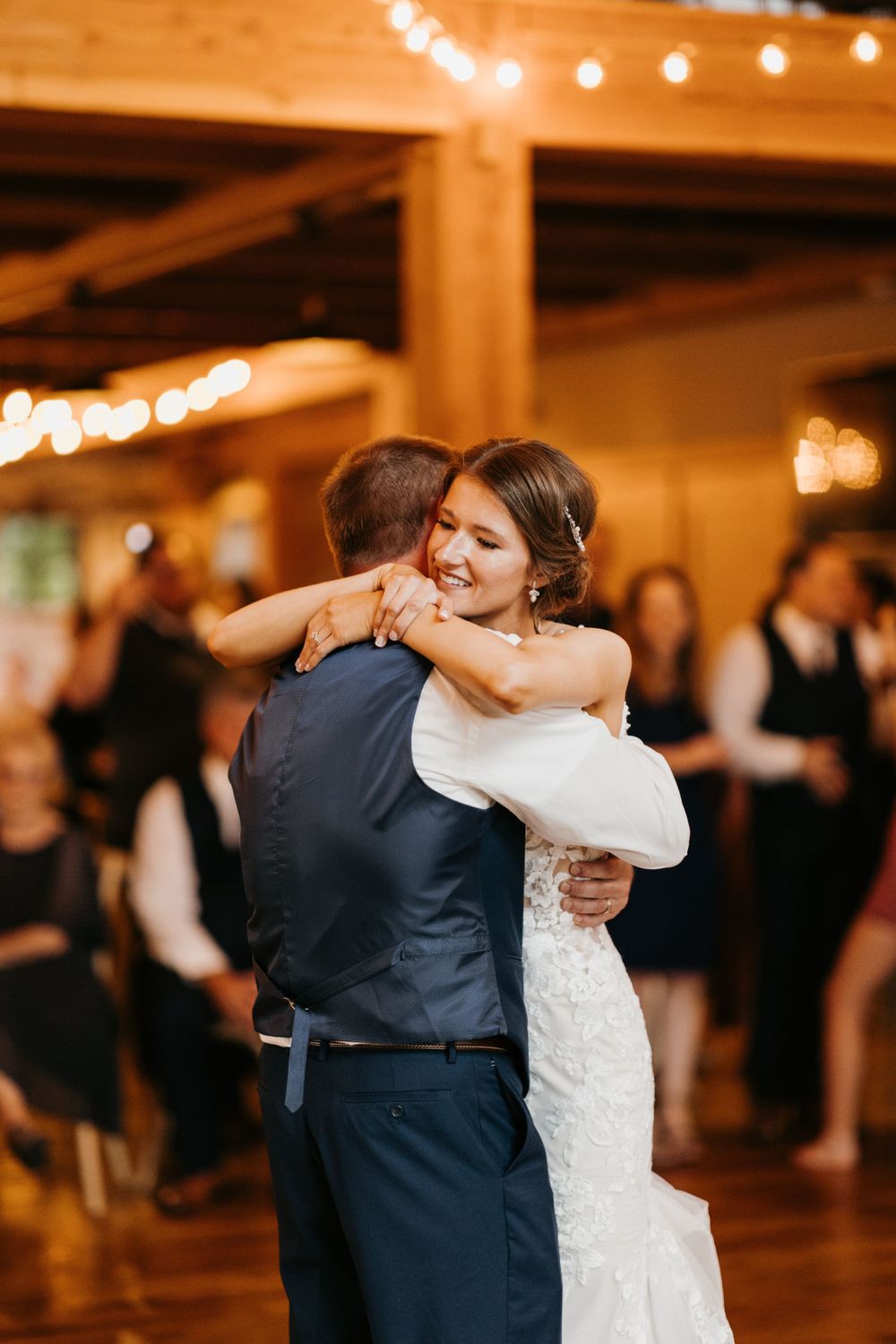 Bride in lace gown smiles while embracing a guest during a warm, glowing barn wedding reception dance.