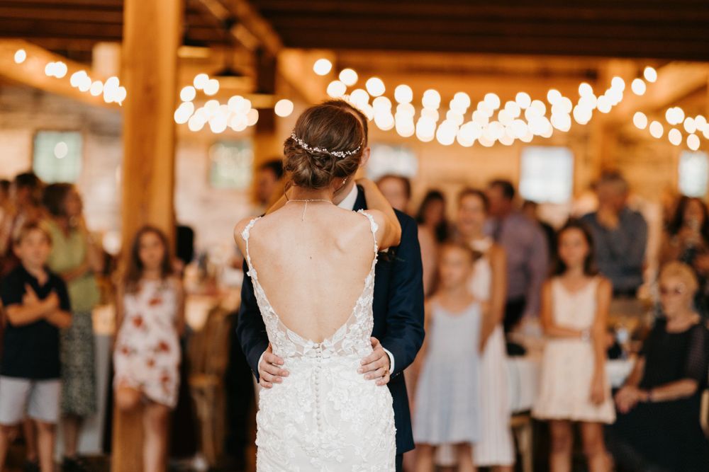 Bride in backless lace gown dances with groom as wedding guests watch in a rustic barn venue.