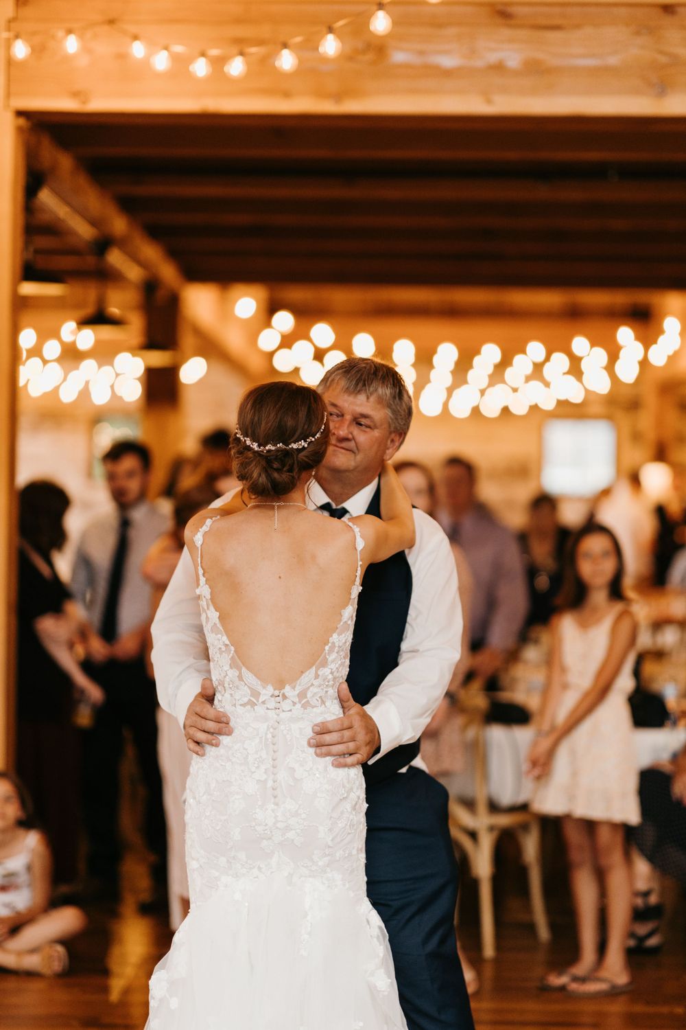 Bride in backless lace gown dances with her father under string lights at a barn wedding reception.