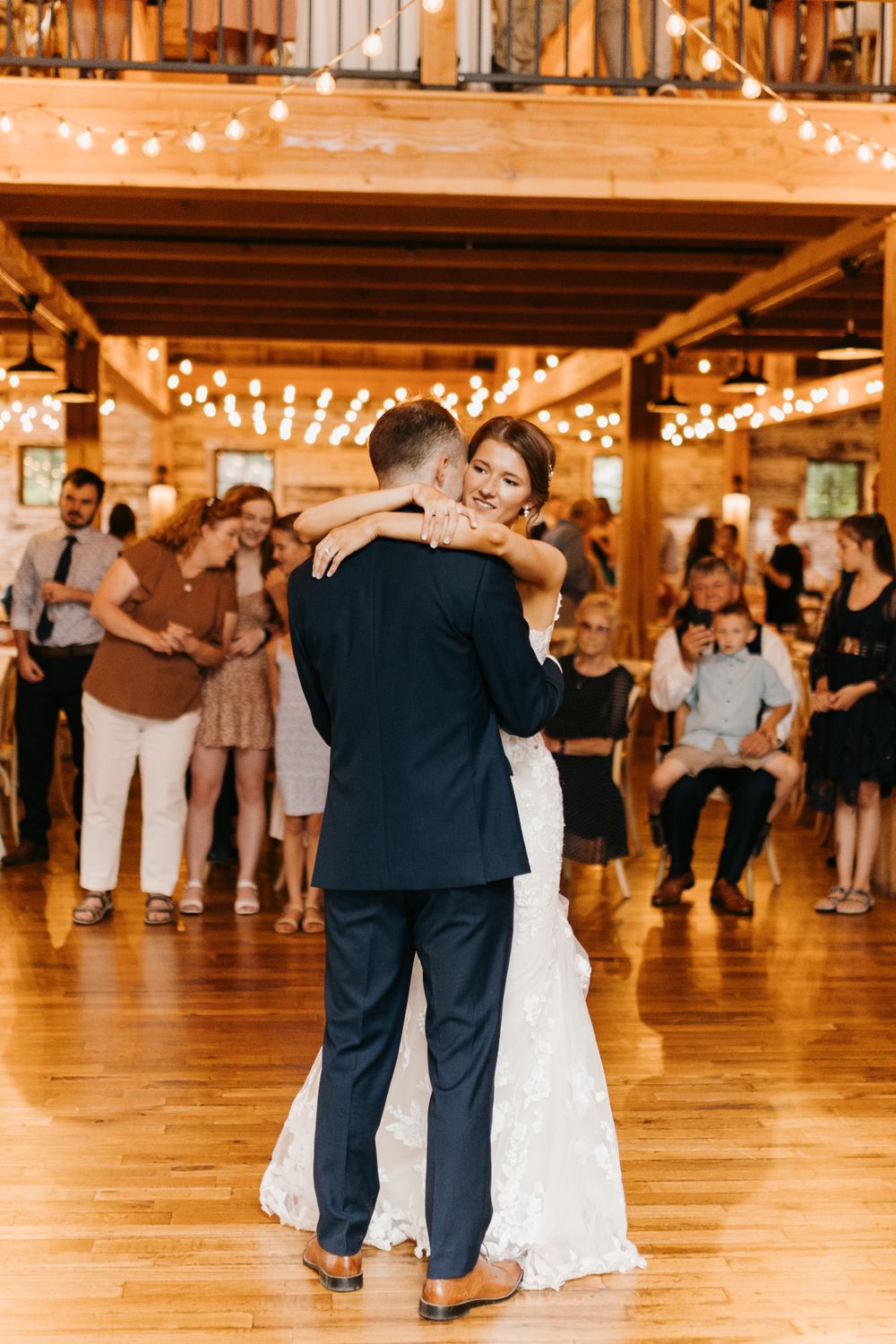 Bride and groom sharing their first dance in a warmly lit rustic barn venue surrounded by wedding guests.