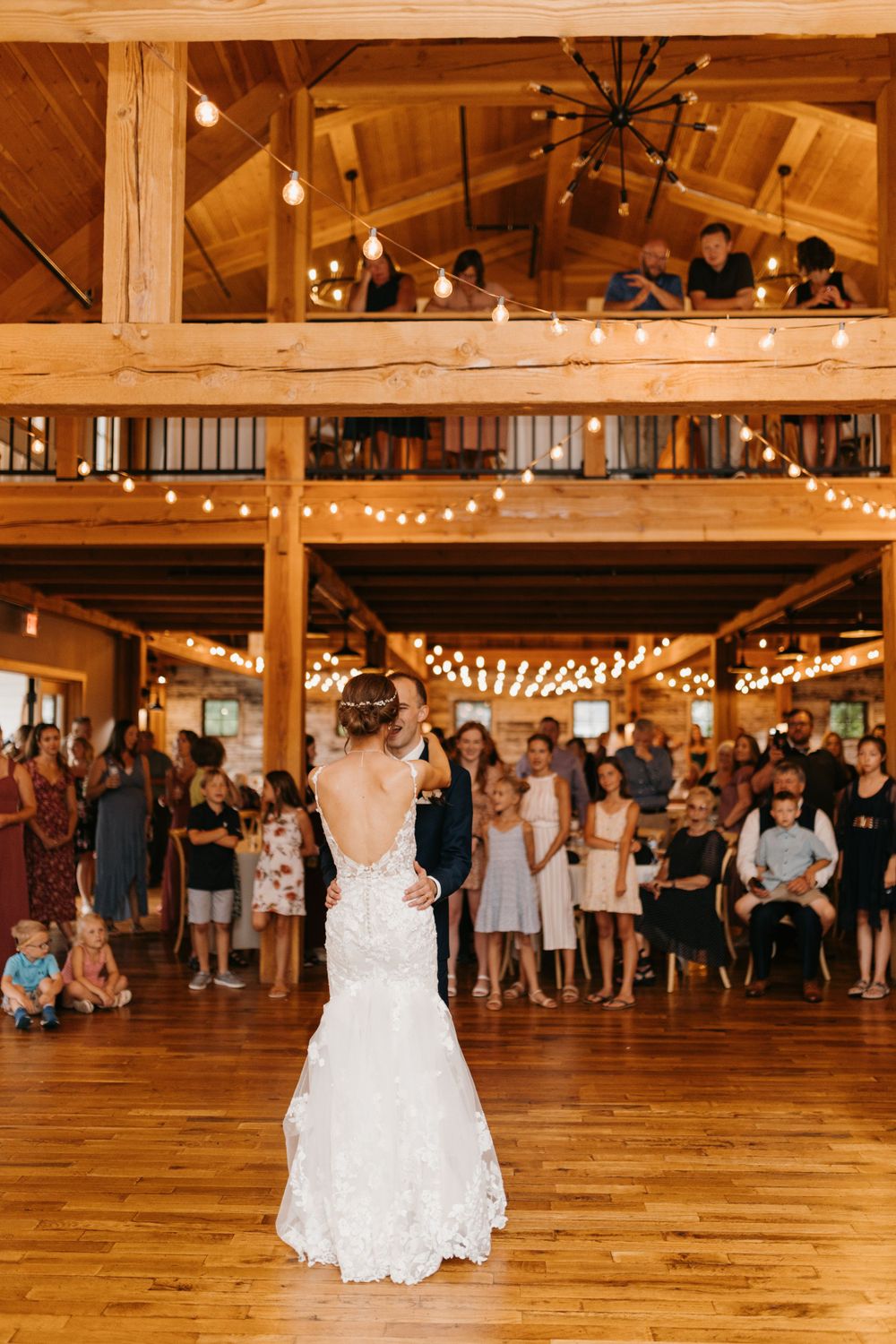 Bride in lace backless gown dancing with groom in rustic barn venue with string lights and guests watching.