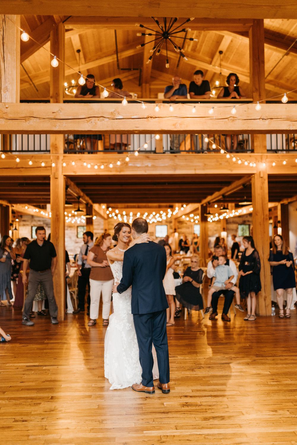 Newlyweds sharing first dance on wooden floor of rustic barn venue with string lights and guests watching.