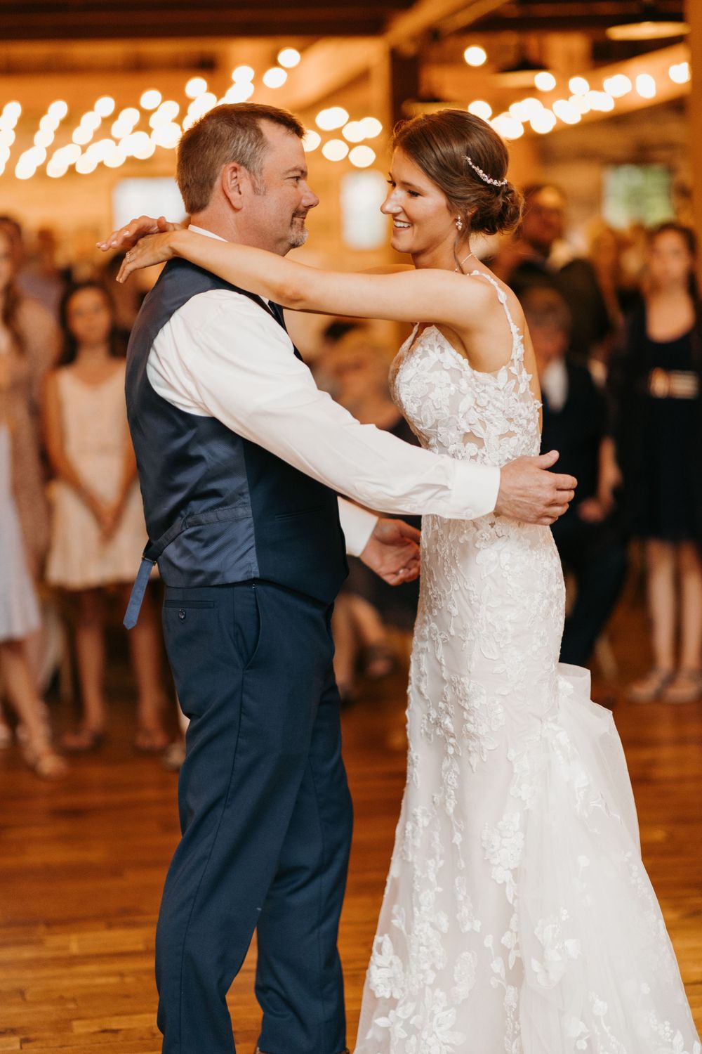 Smiling bride in lace gown dances with her father under string lights at a barn wedding reception.