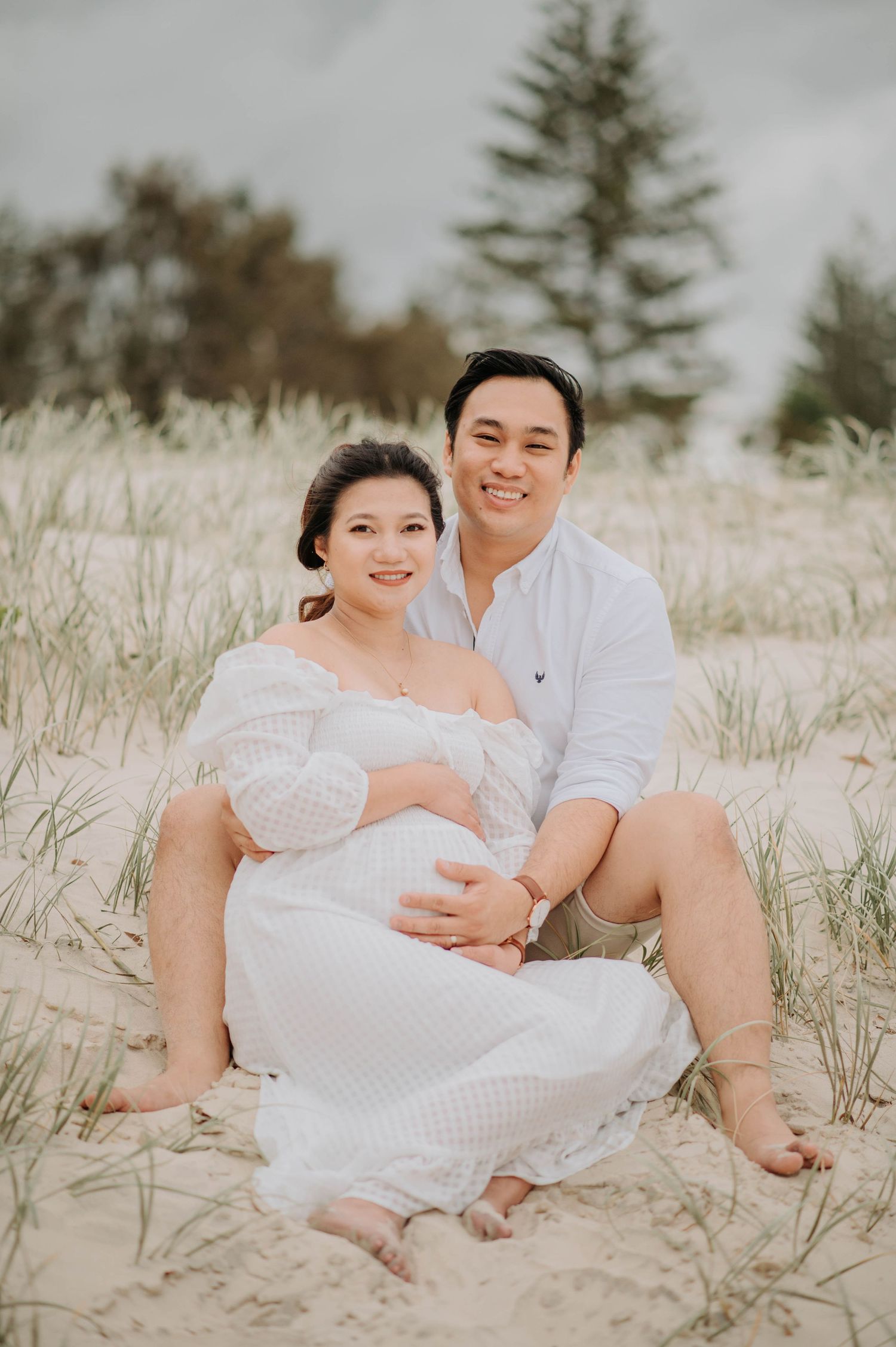 An expectant couple poses together on sandy dunes in white outfits.