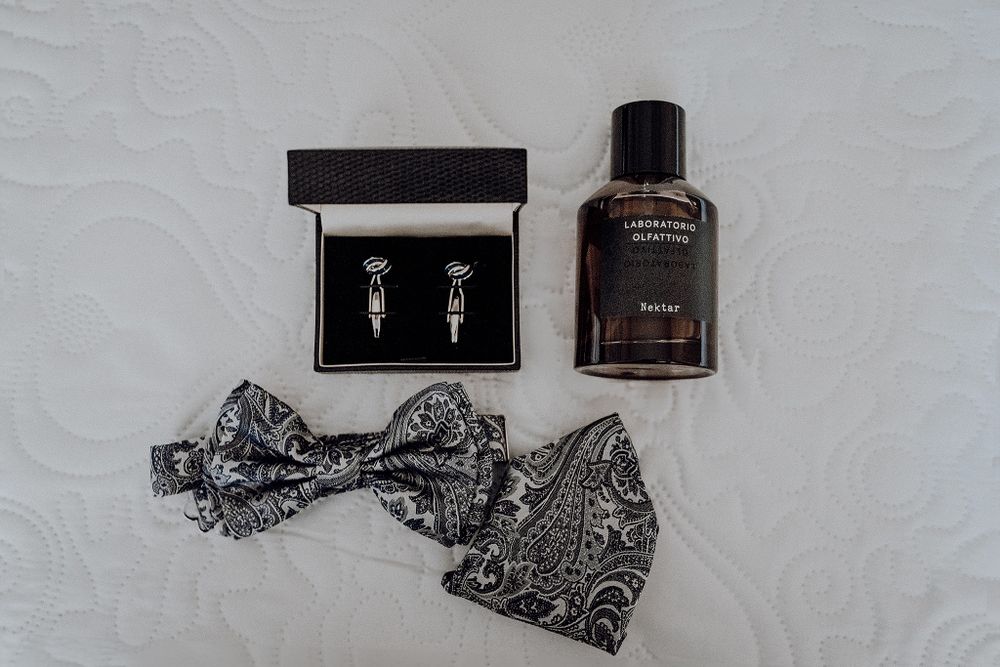 Wedding groom accessories flat lay featuring bow tie, pocket square, cufflinks, and cologne bottle.