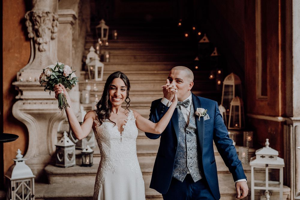 Joyful bride and groom walking hand in hand down ornate stairs, groom kissing her hand at wedding reception.