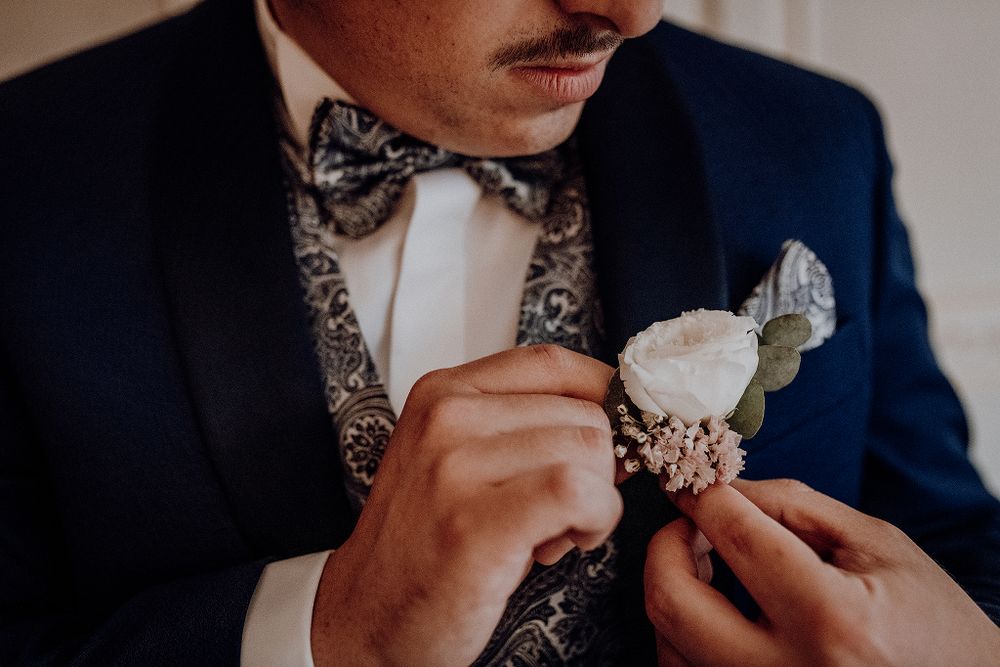 Groom with mustache and floral bow tie pinning white boutonniere on navy suit.