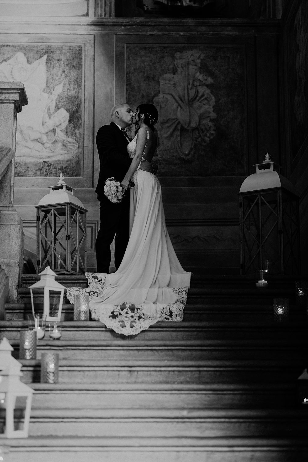 Black and white photo of bride and groom kissing on candlelit stairs adorned with lanterns in ornate venue.
