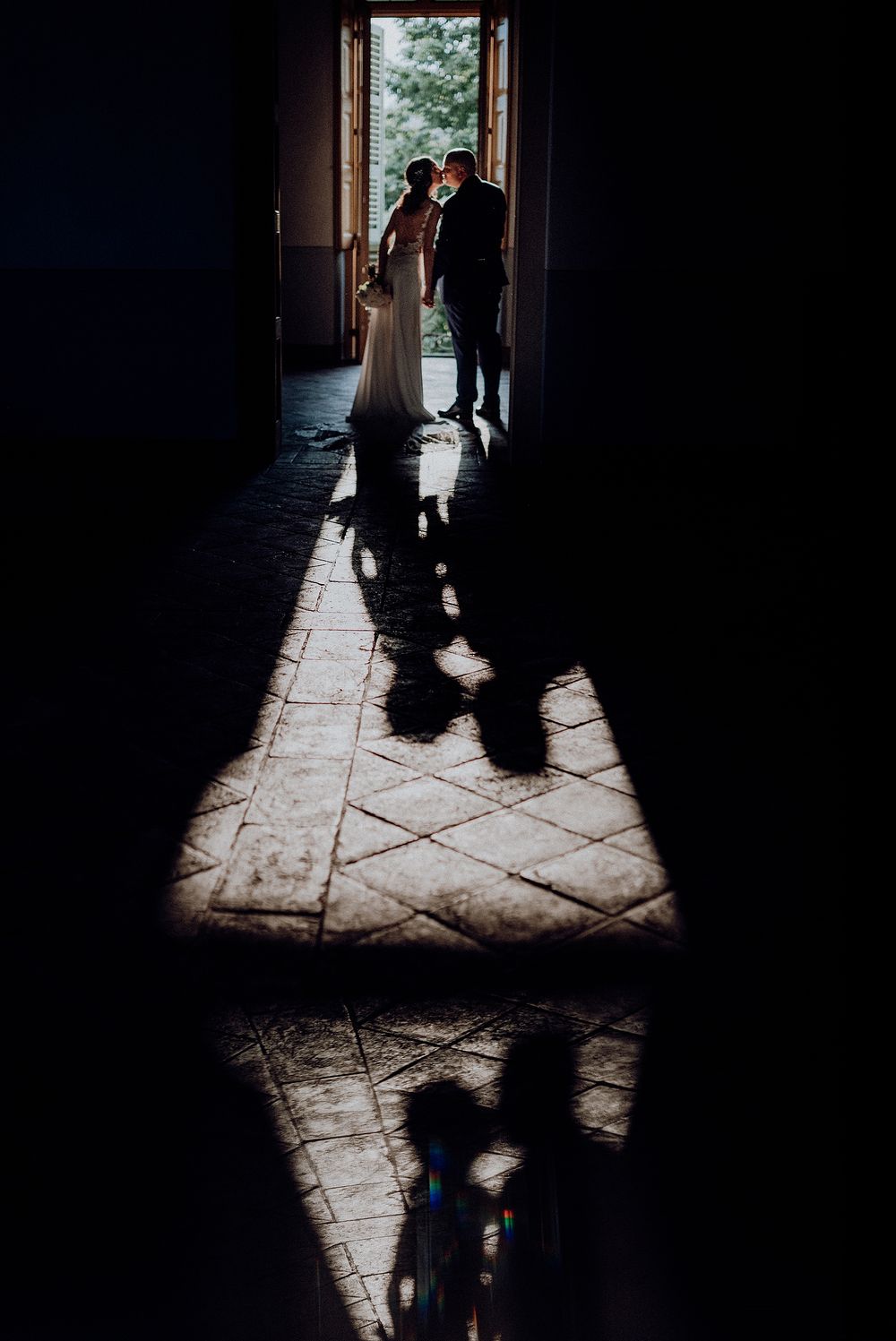 Silhouette of bride and groom kissing in doorway with dramatic shadow reflections on tiled floor.