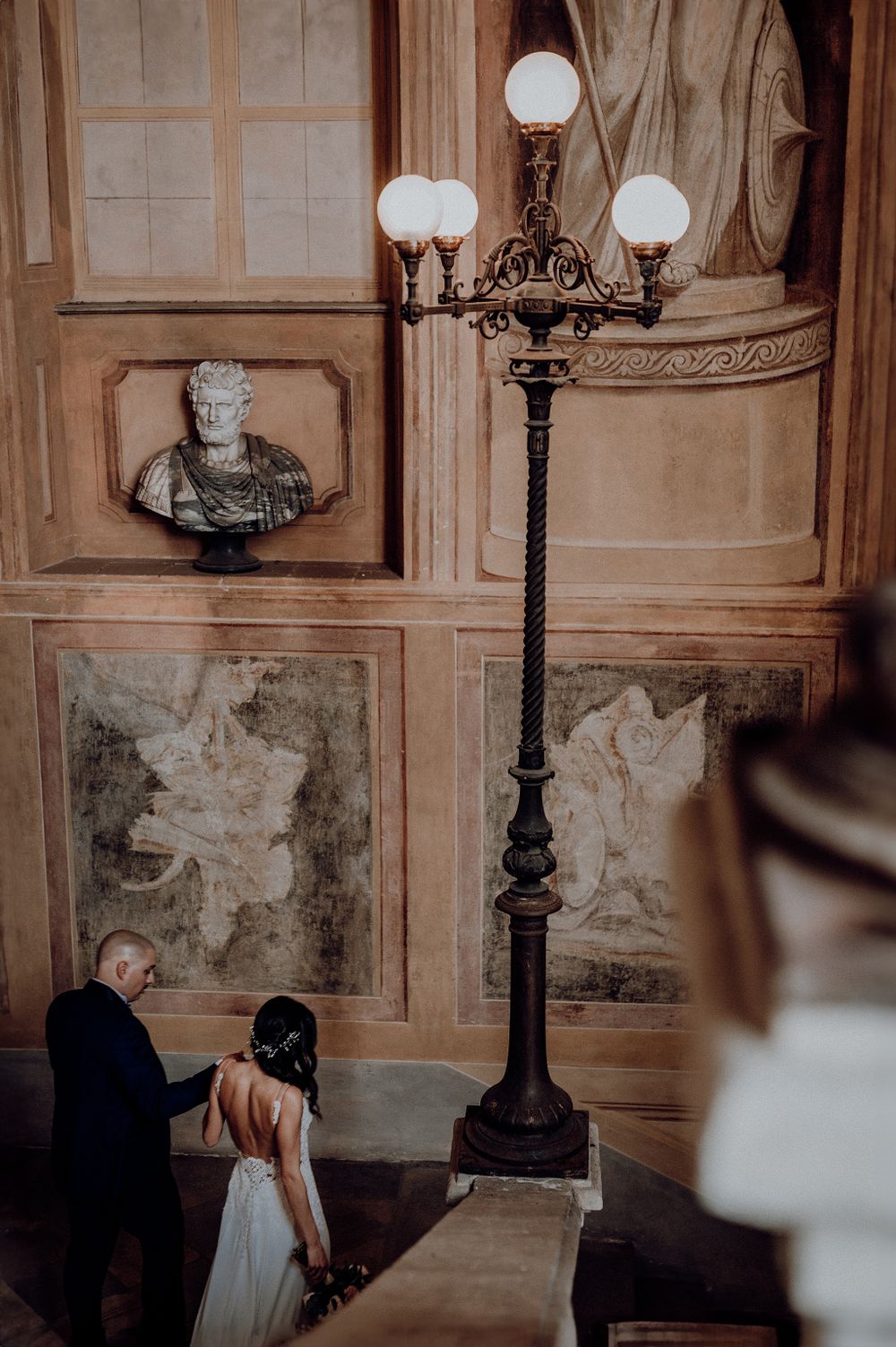 Bride and groom descend ornate staircase in elegant historic venue with classical sculptures and decorative lamp.