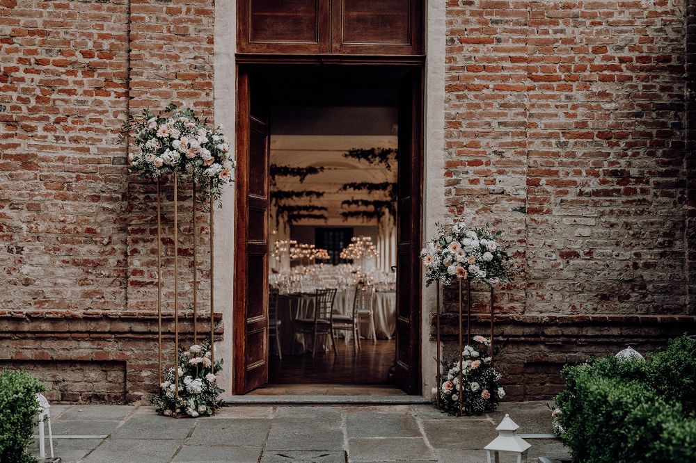 Rustic brick wedding venue entrance adorned with tall floral arrangements flanking grand wooden doors.