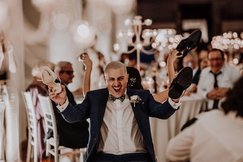 Excited groom celebrating with raised arms and shoes during lively wedding reception.
