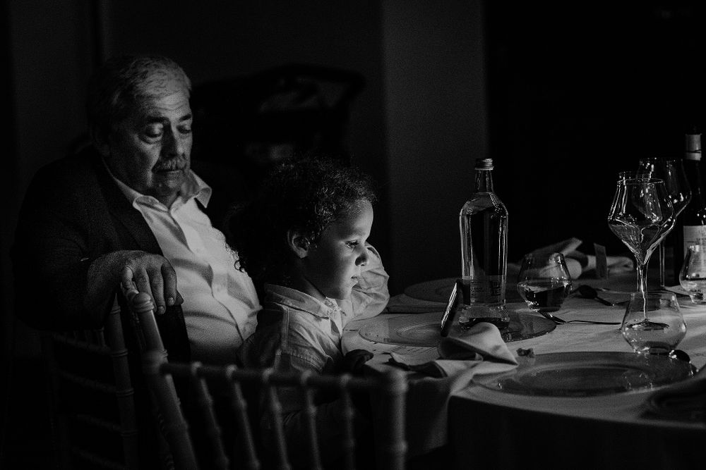Black and white photo of an elderly man and young child sitting together at a formal dinner table with wine glasses.