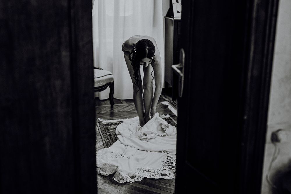 Black and white photo of a bride adjusting her wedding gown in an elegant room.