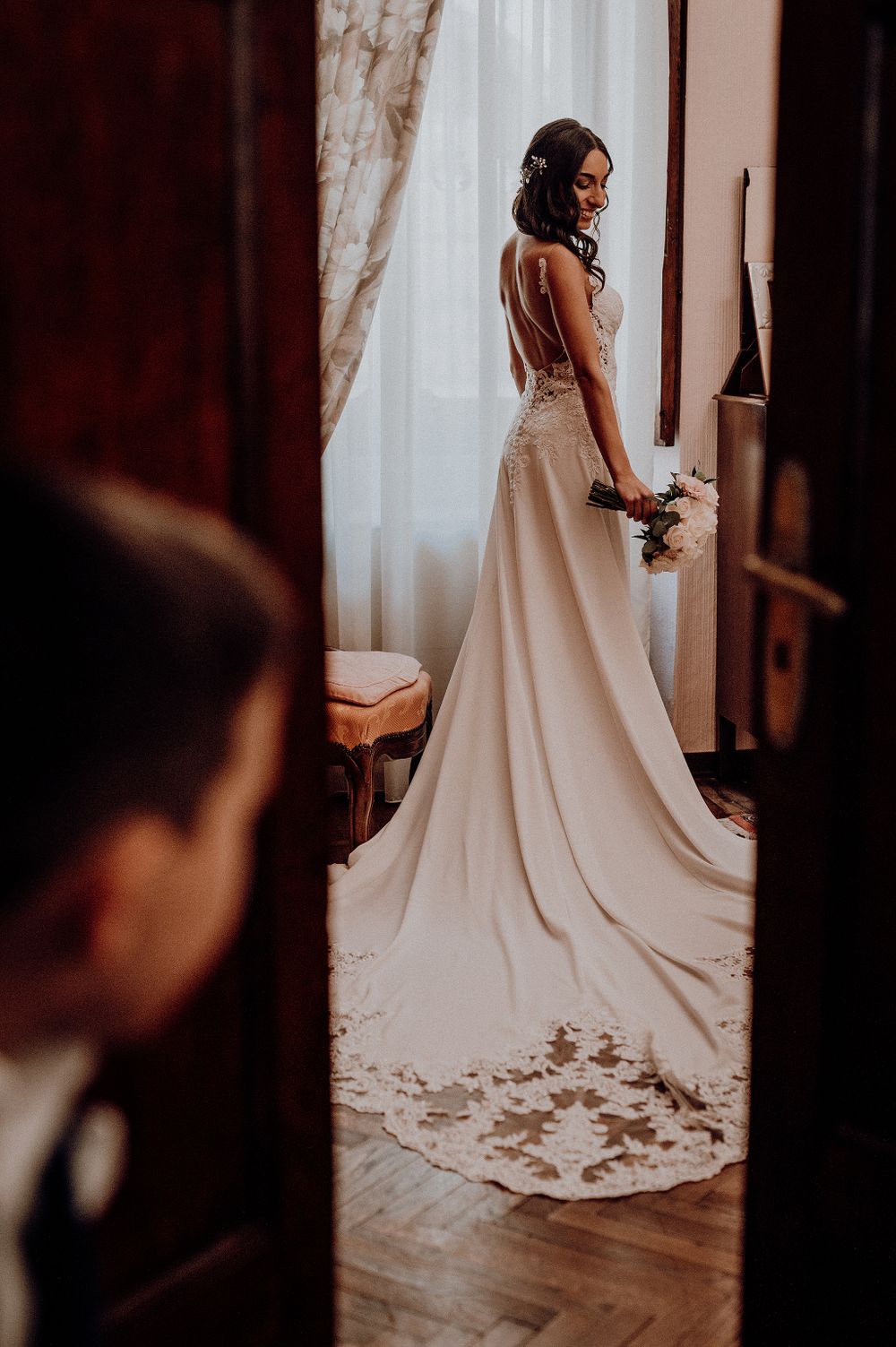 Elegant bride in a lace wedding gown holding a bouquet standing by a window.