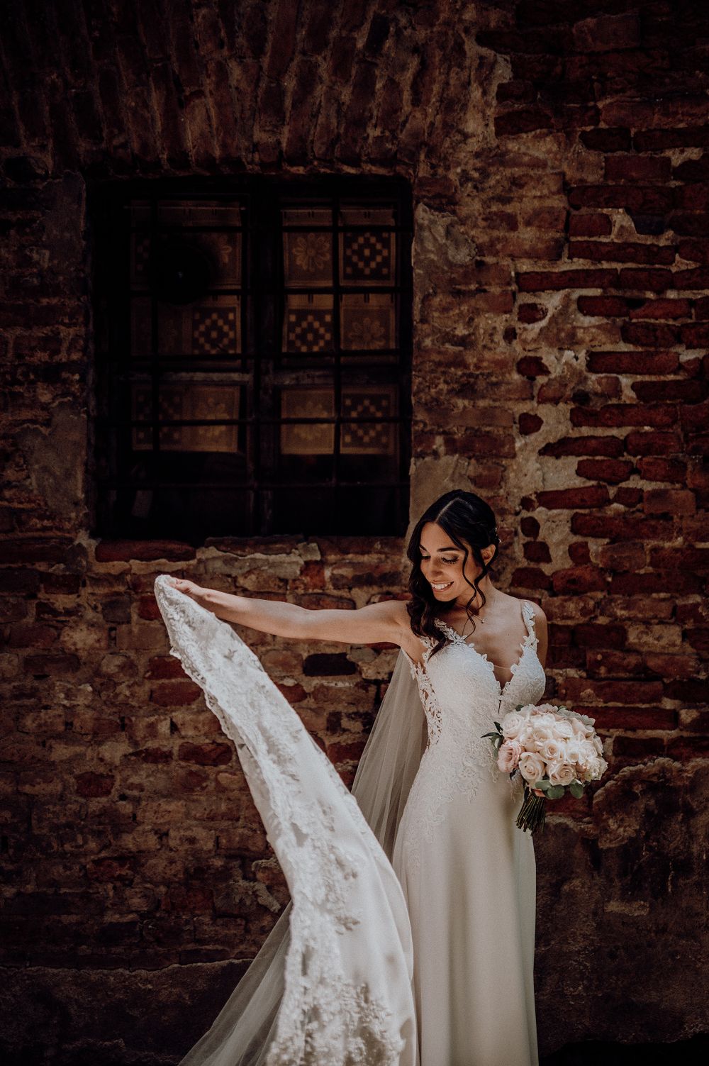 Bride twirling her lace wedding dress veil against a rustic brick wall backdrop.
