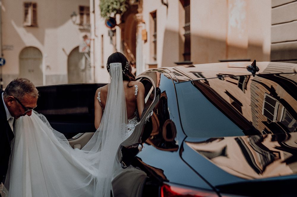 Bride in elegant white gown and veil entering a luxury black car on a charming European cobblestone street.