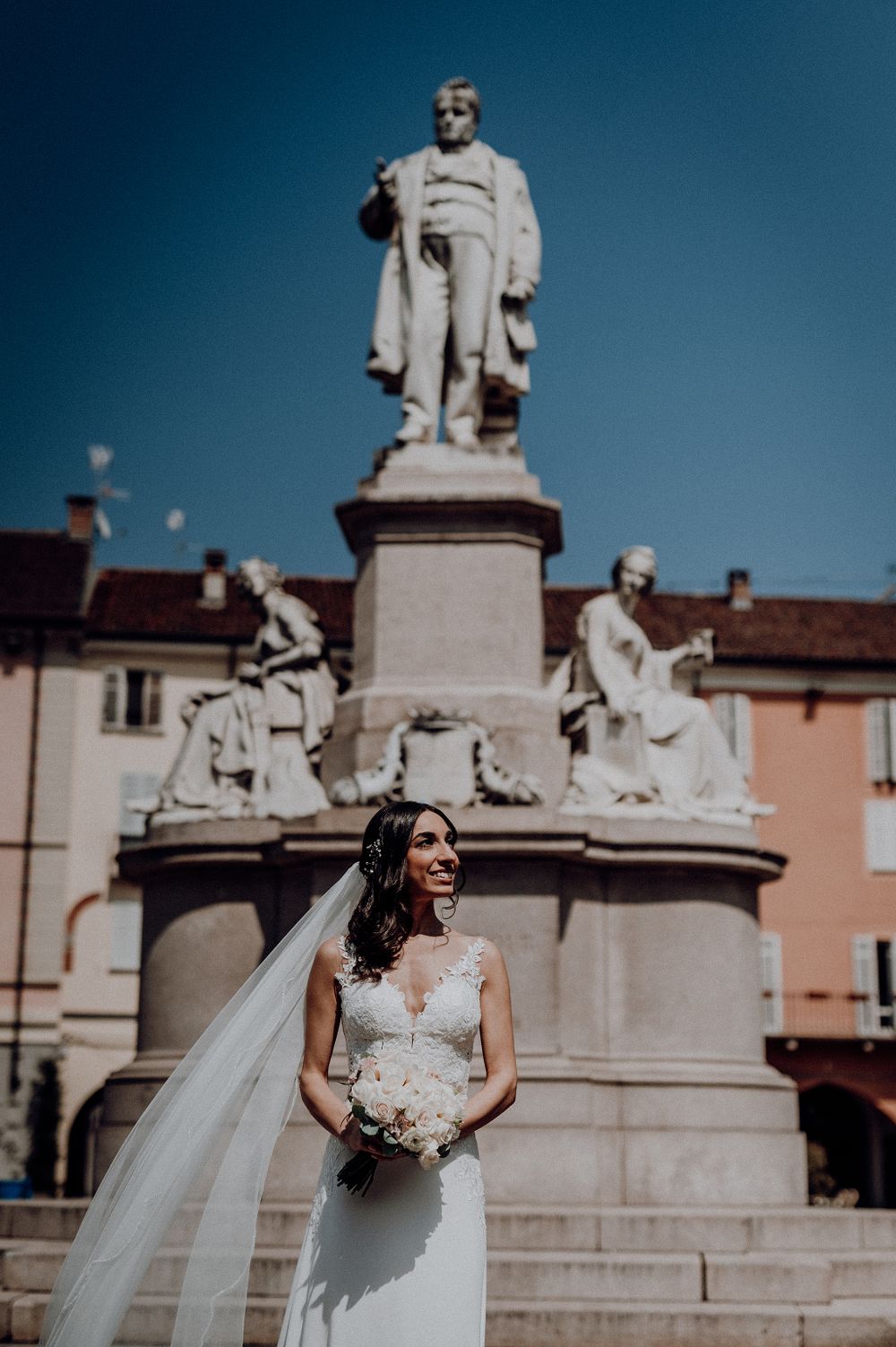 Radiant bride in lace wedding dress poses with flowing veil before a grand classical statue on a sunny day.