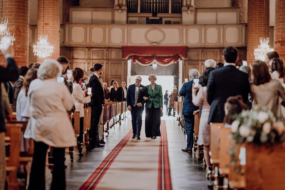 Couple walks down the aisle of an elegant church interior with wooden pews filled with wedding guests.