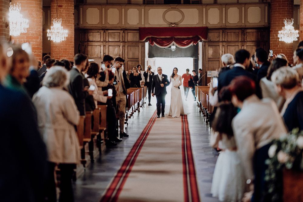 Bride walks down the aisle with escort in a grand church filled with standing wedding guests.