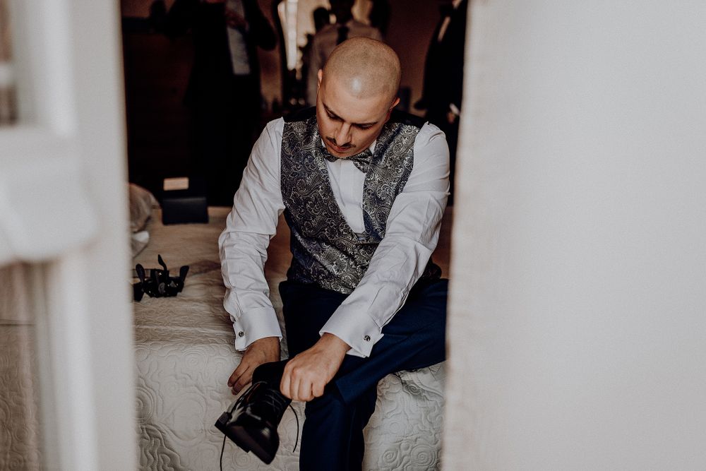 Bald groom in vest and bow tie putting on dress shoes before wedding.