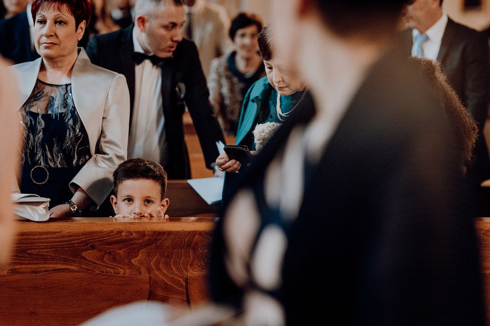 Curious young boy peeks over a wooden church pew during a wedding ceremony surrounded by guests.