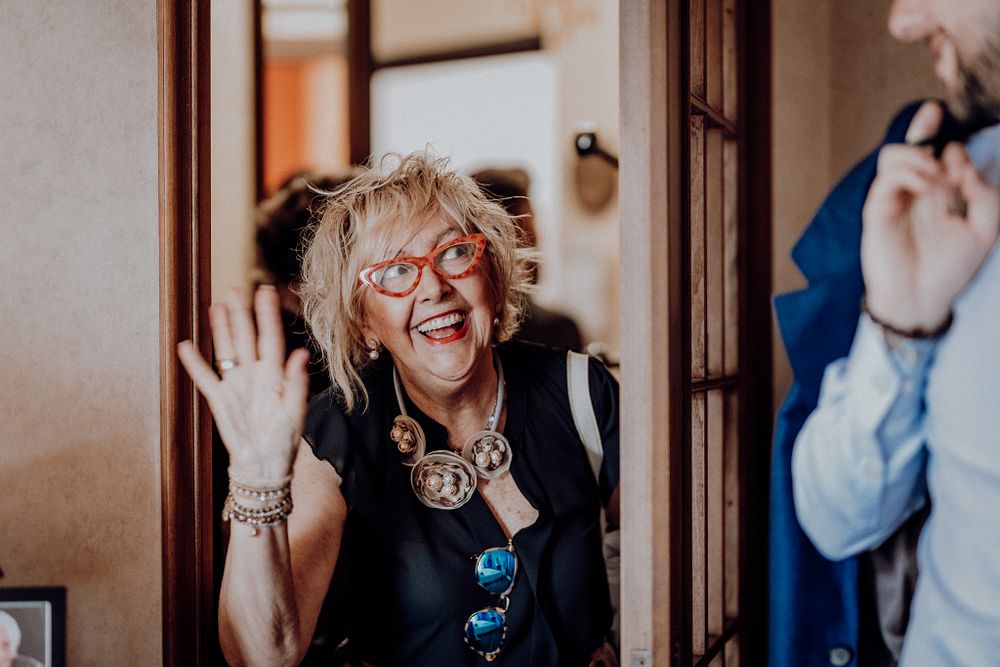 Joyful woman with red glasses and bold jewelry waving at wedding celebration.