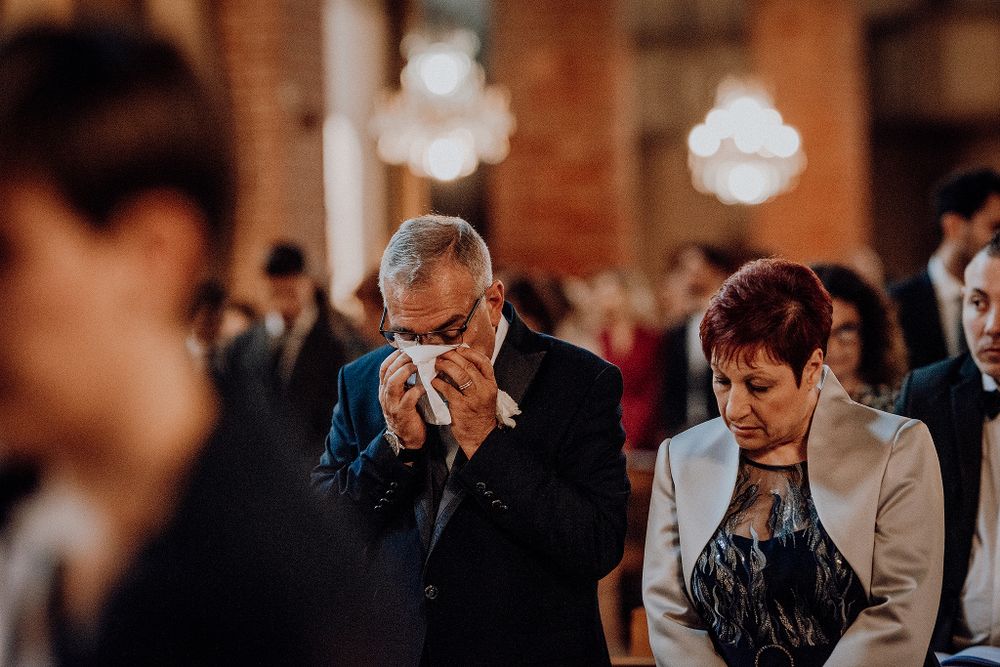 Emotional wedding guest wiping tears during church ceremony beside seated woman in beige jacket.
