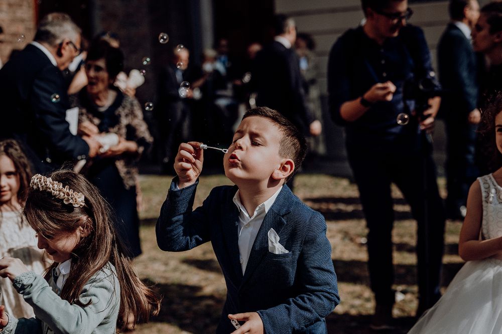 Young boy in blue suit blowing bubbles at outdoor wedding celebration with guests nearby.