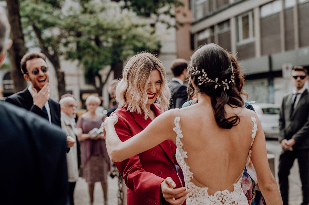 Woman in red wrap adjusting bride's open-back lace wedding dress outside after ceremony.