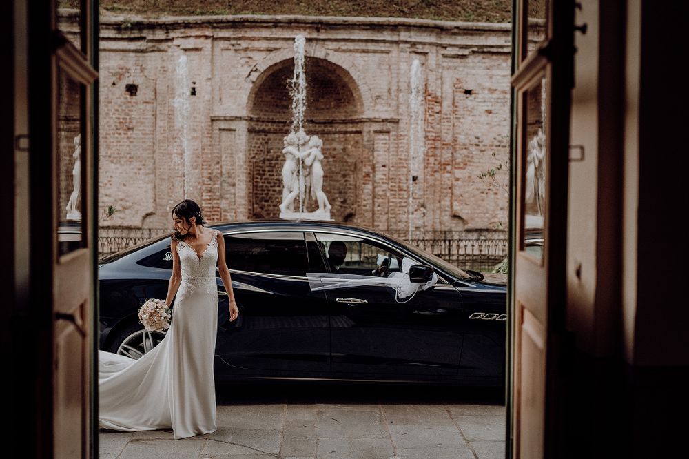 Bride in white gown standing beside black luxury car framed by ornate doorway with fountain.