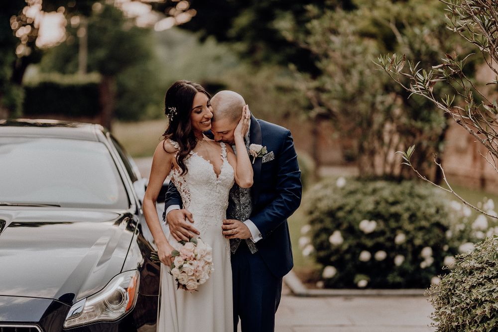 Bride and groom sharing a tender moment beside a black car, surrounded by lush greenery on their wedding day.